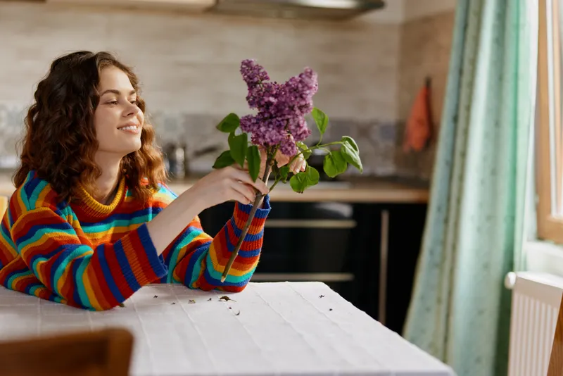 A woman in a colorful striped sweater sits at a bright kitchen table admiring a lilac bouquet, a cozy home moment that captures simple joy, creativity, and mindful atmosphere.