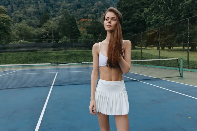 Young athletic female tennis player in a white skirt and top showing confidence on a tennis court surrounded by greenery, embodying a healthy lifestyle