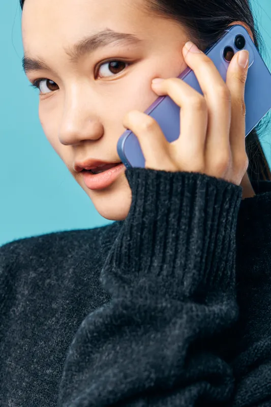 Young woman with smartphone talking on phone, closeup portrait with hand holding device at ear, casual outfit and attentive expression on blue background with natural light indoors