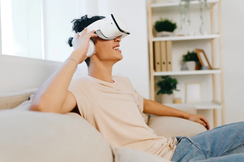 Молодая женщина удобно сидит на диване в VR-очках и улыбается, погружаясь в опыт виртуальной реальности