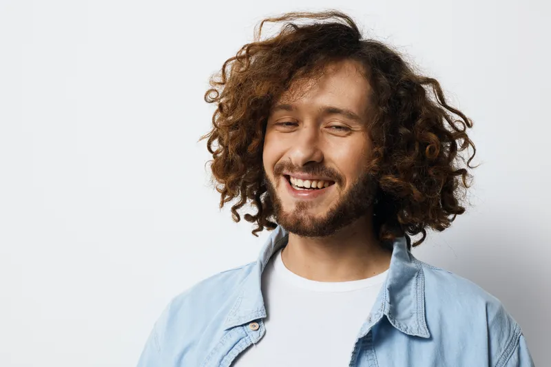 Happy young man with curly hair wearing a light blue shirt smiling against a neutral background, representing joy, positivity, and confidence in casual fashion