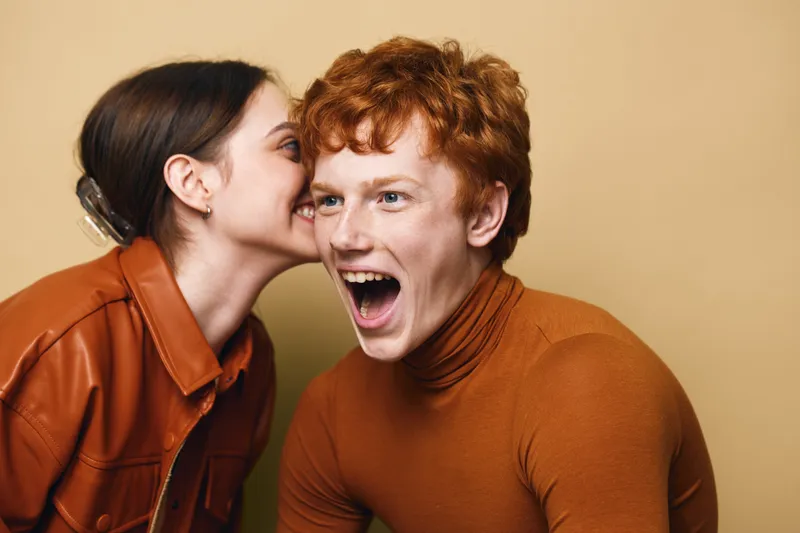 Young woman whispering secret to surprised young man with red hair against beige background. Casual clothing and emotional facial expressions in portrait studio shot.
