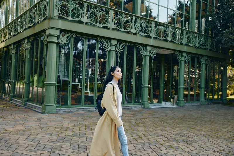 Woman with backpack in a long beige coat strolling beside an ornate glass and metal structure, outdoor courtyard setting, architectural detail, fashion mood, candid street scene