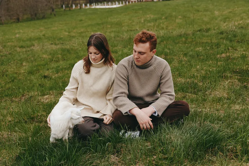 Two people sit close on a grassy field in knit sweaters, enjoying a calm outdoor moment, natural light, and a warm lifestyle mood, capturing intimate togetherness and quiet happiness