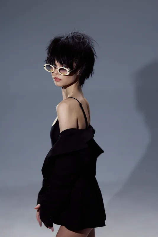 Stylish woman with short hair and sunglasses wearing a black outfit posing in a minimalist studio setting.