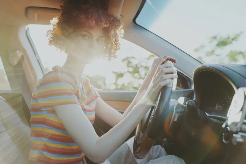 Cheerful woman driver in a sunlit car, striped shirt and curly hair, relaxed lifestyle moment, warm retro color, natural candid atmosphere