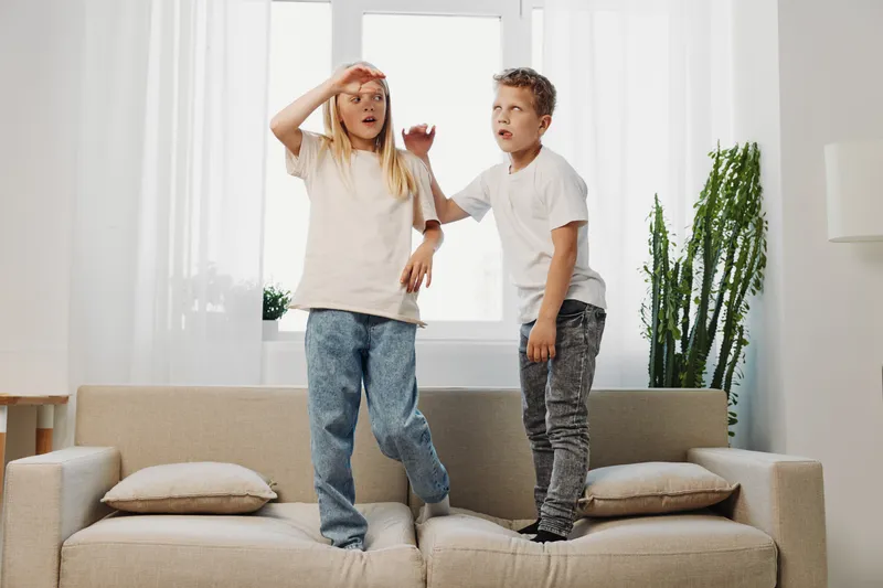 Two children stand on a sofa in a bright living room, expressing surprise and playfulness, capturing a lively moment of fun and interaction at home.