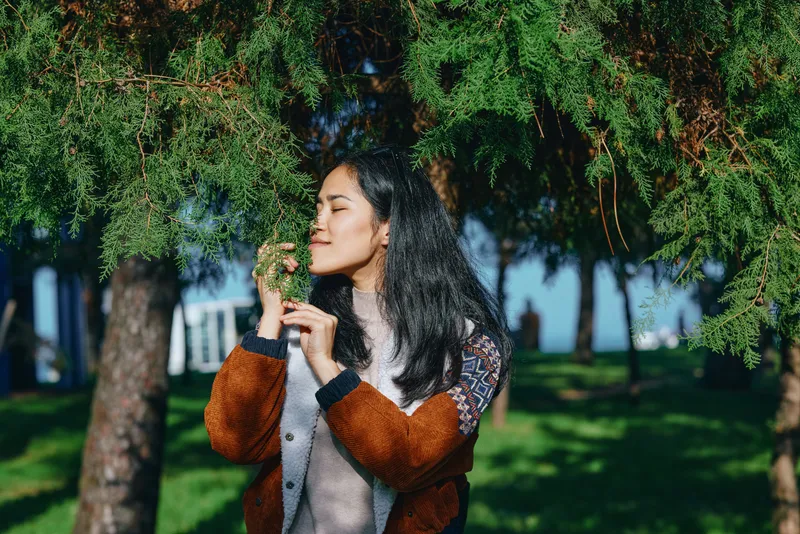 Woman enjoying nature in a park, smelling leaves among lush greenery, serene moment outdoors, casual jacket, dappled light, tranquil mood, fresh air, seasonal atmosphere