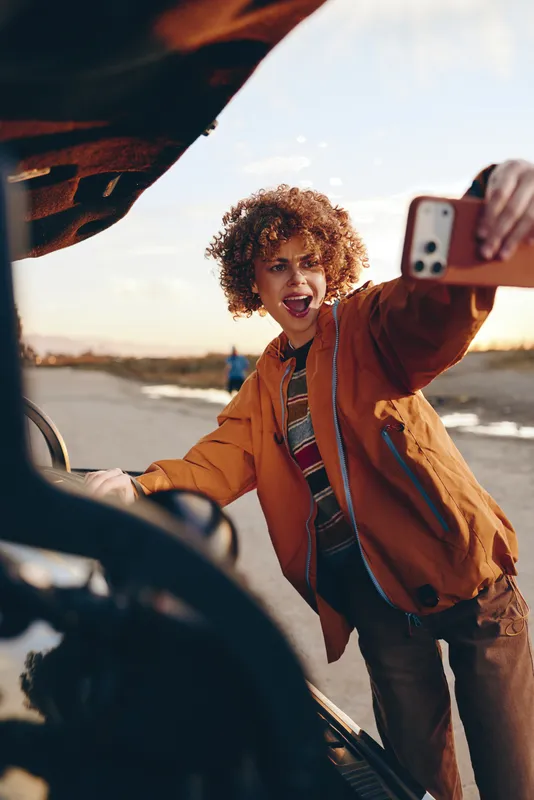 Curly haired woman outdoors smiling and taking a selfie near a parked car, warm sunshine, casual travel lifestyle, autumn jacket, open road background, relaxed pose, golden hour light