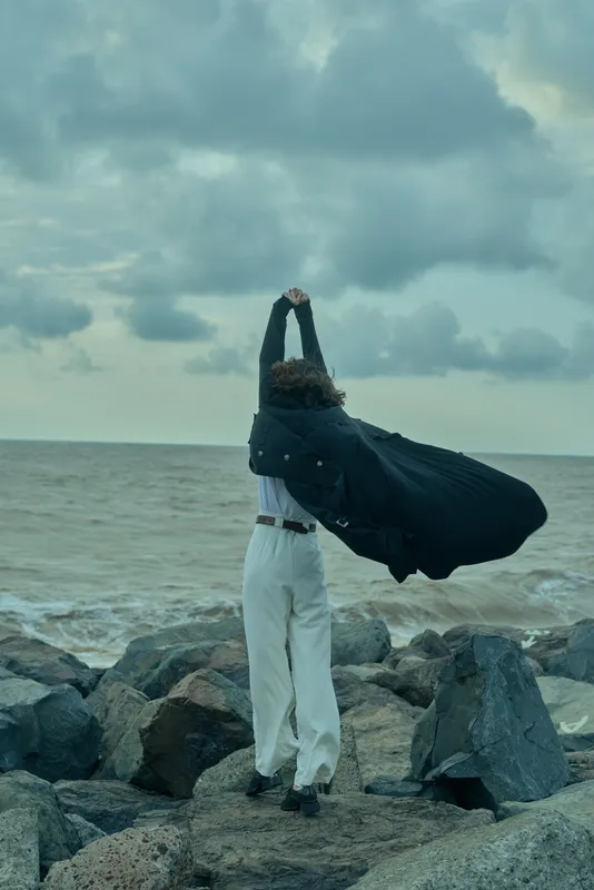 Woman on a rocky shore, cape flowing as she stretches arms to the sky, windy sea scene conveying freedom, outdoor adventure, and resilience