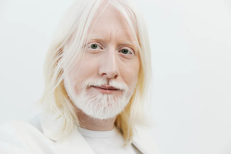 На этом изображении изображен пожилой мужчина с длинными седыми волосами и бородой, одетый в белый костюм