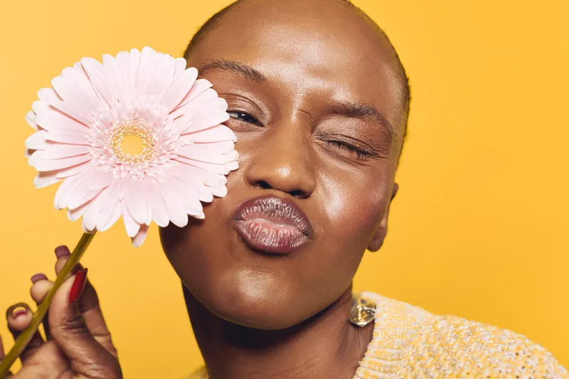 Close-up of a happy dark skinned African woman with smooth skin holding a pink flower near her face. She makes a playful kiss expression on a bright yellow background. Concept of beauty and joy.