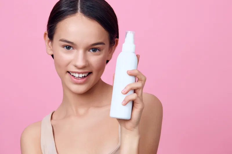 Young woman with a natural glow holding a white skincare bottle against a pink background, symbolizing beauty and skincare concepts