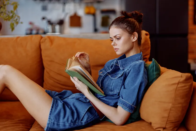 Молодая женщина в джинсовом платье читает книгу, сидя на диване в уютной гостиной у себя дома
