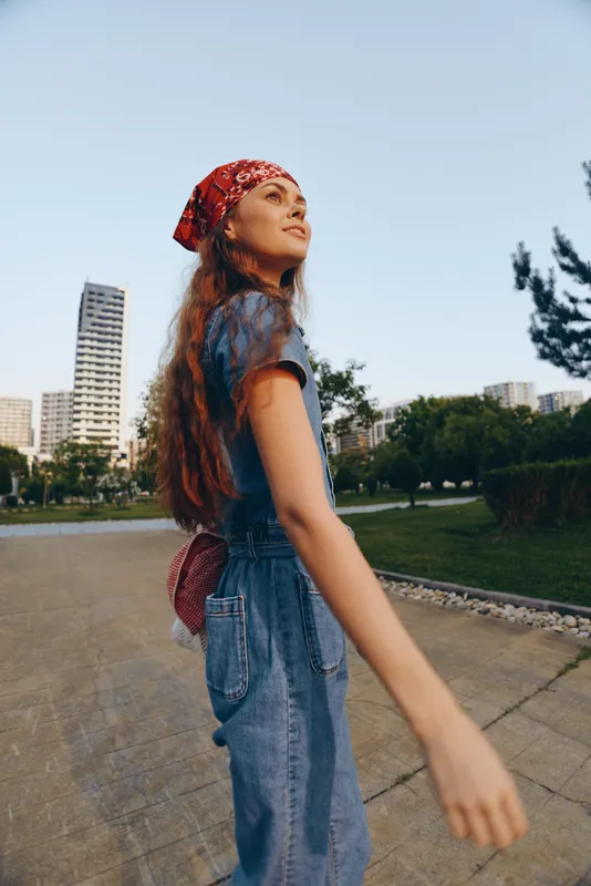 Young woman wearing blue denim overalls and red bandana walking outdoors in an urban park with modern buildings and green trees in the background.