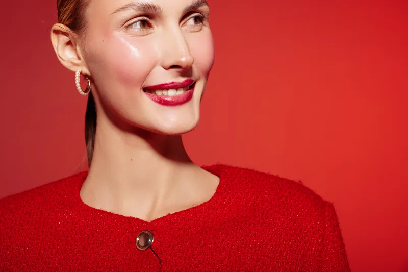 Smiling woman in red sweater looks away on red background. Close-up portrait with natural makeup and hoop earrings. Concept of happiness, fashion, and beauty in studio lighting.