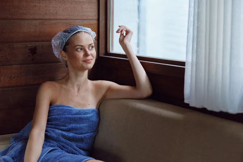 Relaxed woman in spa with towel and shower cap sitting by window, enjoying peaceful moment in cozy wooden interior, soft natural light highlighting calm expression.
