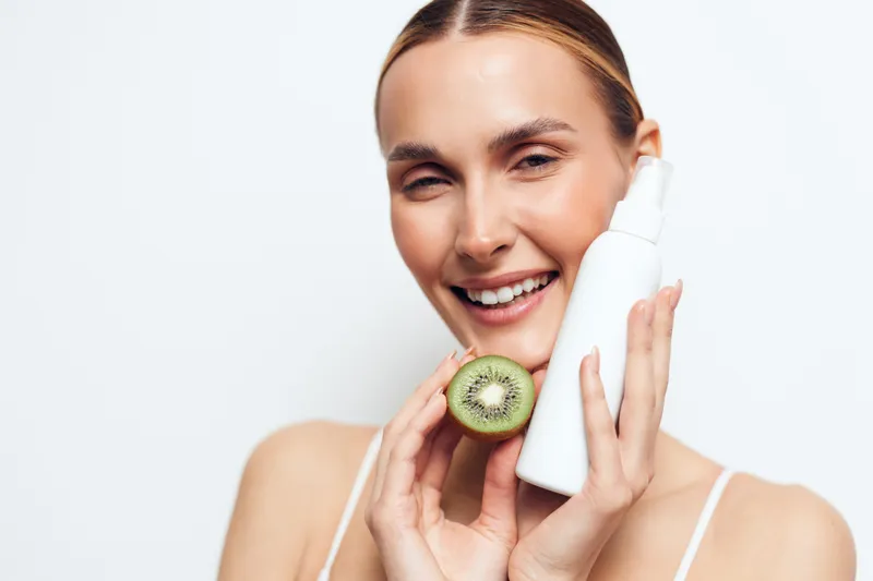 Young woman with blonde hair smiles while holding a white cosmetic bottle and a fresh kiwi slice against a white background in a skincare beauty concept.