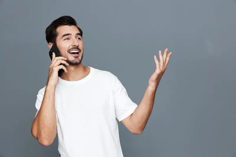 Smiling man talking on mobile phone, casual white t-shirt, expressing happiness and excitement, isolated on plain grey background, studio shot. People lifestyle concept