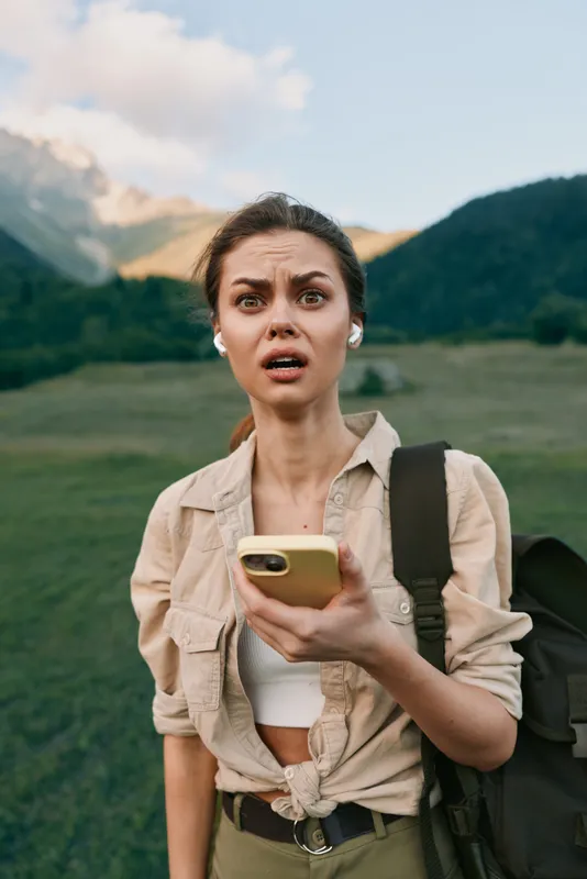 Woman traveler outdoors with backpack holds smartphone, surprised expression, mountain meadow backdrop, casual rugged style, candid lifestyle moment