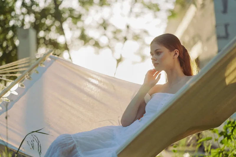 Serene young woman in a flowing white dress, resting on a hammock, enjoying summer sunshine amidst greenery, embodying tranquility and relaxation