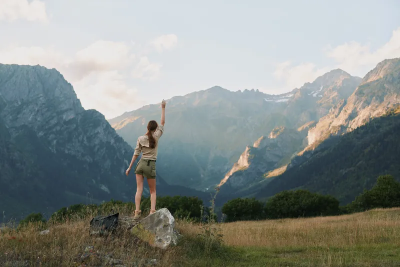 A young woman stands on a rock in a mountain valley, raising her arm in triumph against distant peaks and an open sky, conveying freedom and outdoor adventure.
