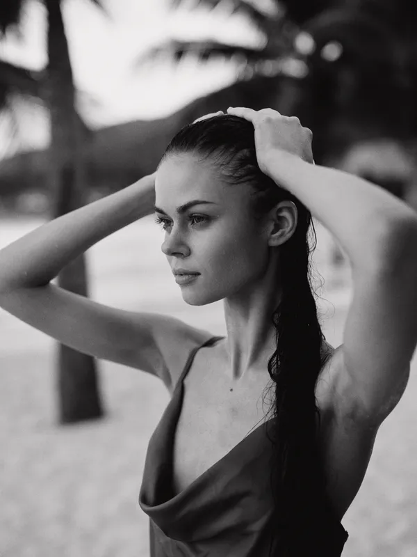 Monochrome portrait of a woman at the beach, hair tied back, hands lifting a towel. Soft light highlights calm features and a contemplative mood, with a graceful silhouette against blurred palm trees