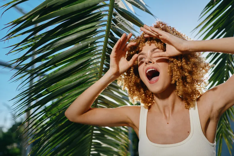 joyful woman with curly hair enjoying sunlight outdoors near palm leaves, wearing white tank top, summer vibes and natural beauty in warm daylight.