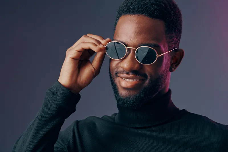 Confident young Black man wearing sunglasses and a turtleneck, smiling against a dark, subtly lit background, representing style and modern fashion