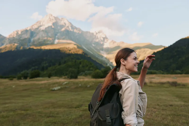Smiling woman hiking outdoors with backpack in mountain landscape, clear sky, natural light, casual clothing, enjoying nature, active lifestyle, fresh air