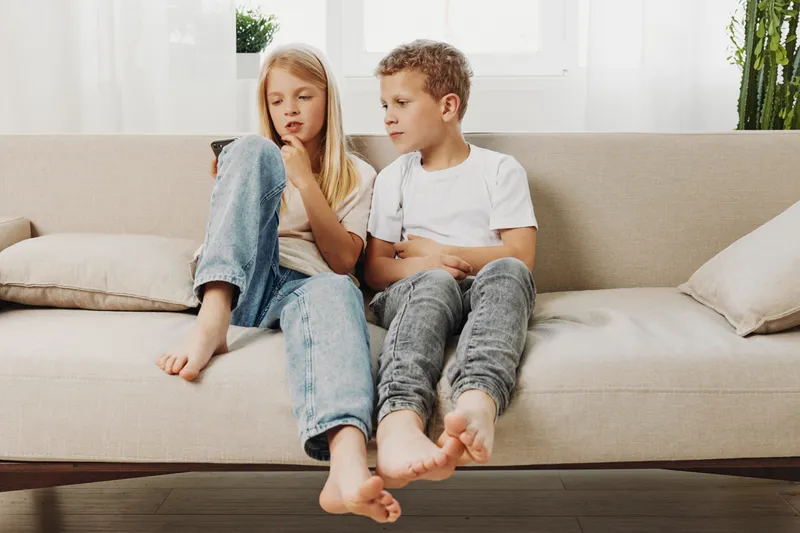 Two children sitting barefoot on beige sofa in casual clothes, focused on smartphone screen in bright living room. Concept of childhood, technology, sibling interaction, and modern family life.
