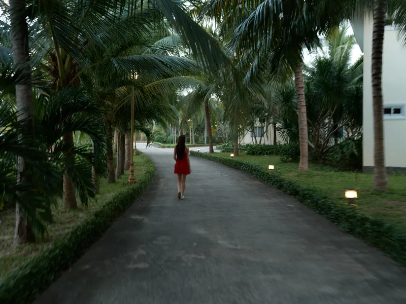 Woman walking in red dress along palm tree-lined path at dusk, tropical atmosphere, calm mood, outdoor lifestyle, serene environment.