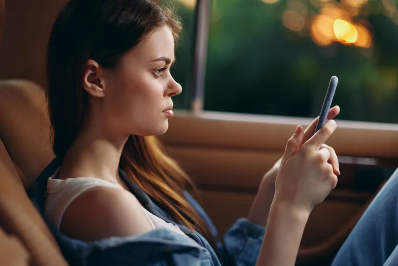 Young woman using smartphone in car during sunset, wearing casual denim clothes, expressing calmness and relaxation in a warm-toned backdrop.