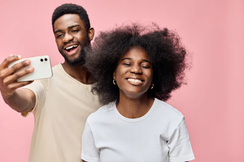 Happy black couple taking selfies together against a pink background, showcasing their joyful emotions, casual outfits, and playful connection
