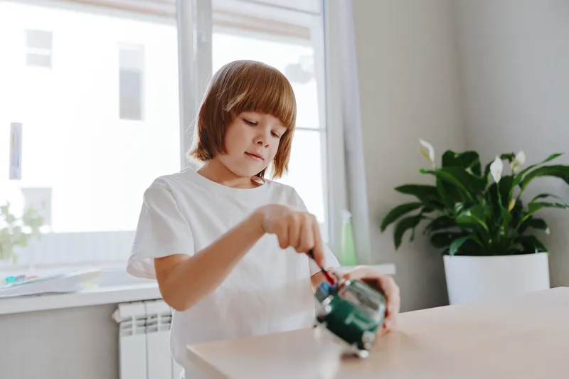 A focused boy is using a screwdriver to work on a small toy. The bright space features plants, creating a productive and lively atmosphere for creativity.