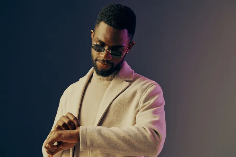 Stylish young Black man in sunglasses looking at his watch, dressed in a beige coat against a gradient background, conveying confidence and elegance