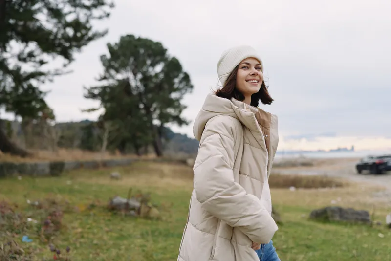 Woman in a beige puffer coat and white beanie outdoors, smiling, standing in a grassy open field with trees and a distant road, bright seasonal portrait