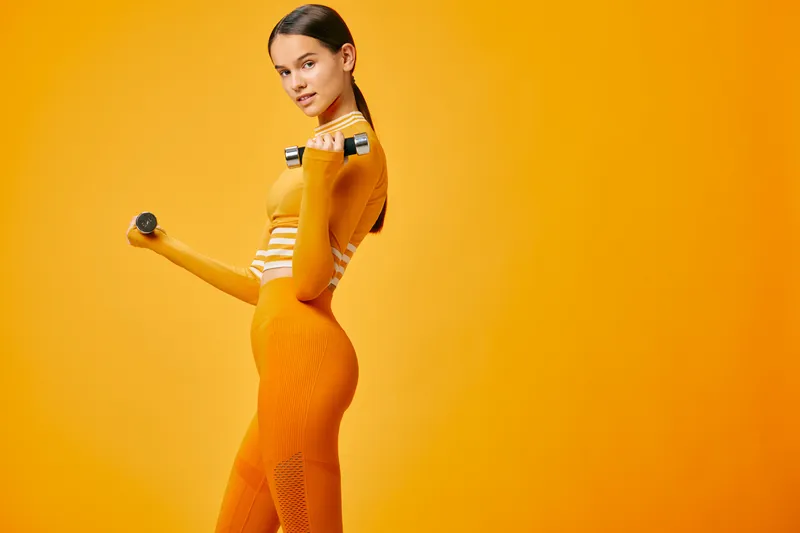 Fit young woman in vibrant orange fitness outfit holds small dumbbells in both hands, posing in studio against solid orange background, braided hair, confident athletic posture.