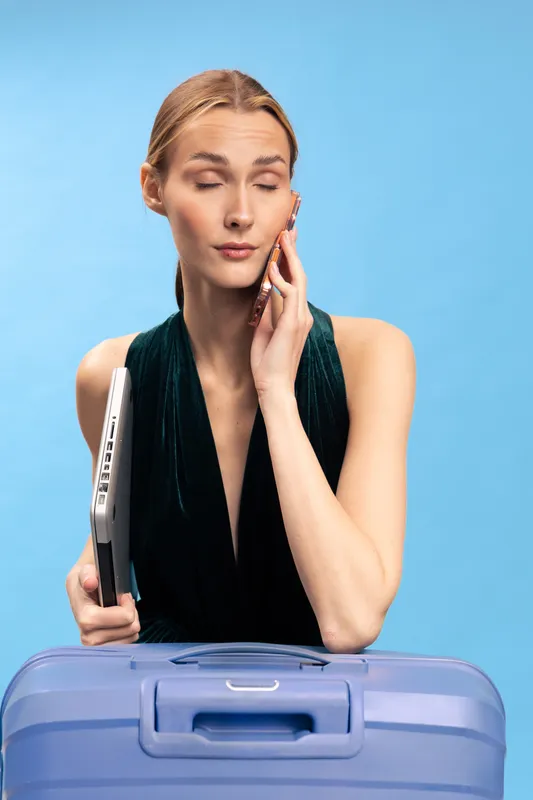 Young woman in black dress holding laptop and talking on phone with eyes closed, standing behind blue suitcase on blue background. Concept of travel, communication, and business trip.