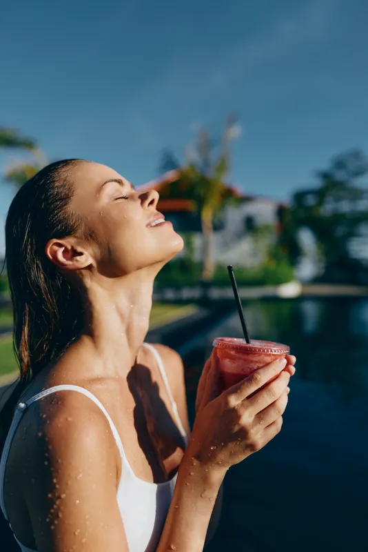 Cheerful woman outdoors by a pool, sunlit moment, sipping a red smoothie, eyes closed in bliss, white swimsuit straps, tropical backdrop, relaxed lifestyle