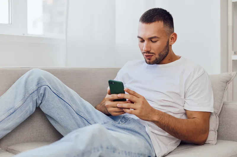 Focused man using smartphone while relaxing on the couch, showcasing modern technology and lifestyle trends. A calm atmosphere in a cozy living space.