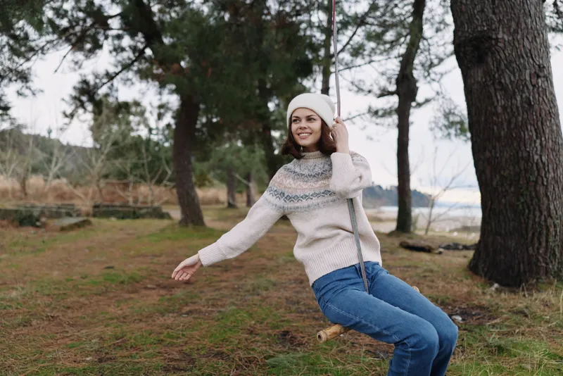 Woman on a swing in an outdoor park, wearing a knit sweater and beanie, smiling as she enjoys a breeze among tall trees and soft grass, a casual moment of nature and freedom.