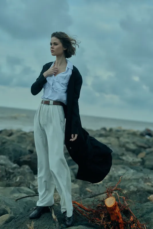 Fashionable woman on a rocky coast, wind in her coat, white shirt and wide trousers, dramatic sea backdrop emphasizing style and resilience