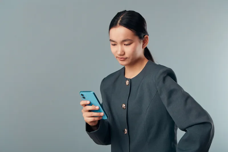 Young woman stands looking at smartphone screen with focus wearing dark jacket against gray background indoor technology communication digital device and casual style.