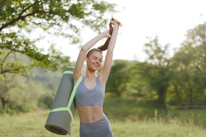 Smiling young woman in activewear holding a yoga mat outdoors, celebrating health and fitness in a serene natural setting