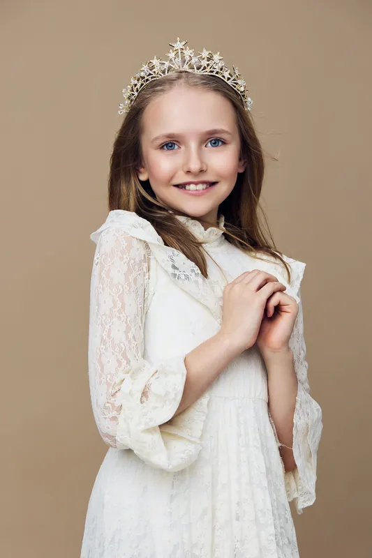 Young girl with a crown smiling against a soft beige background, wearing a delicate white dress, conveying joy and elegance, ideal for children s portraits