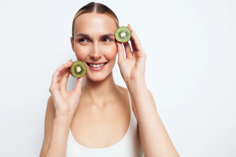 A smiling young woman holds two halves of a kiwi near her face against a white background. The healthy fruit and natural beauty concept emphasizes freshness and wellness.