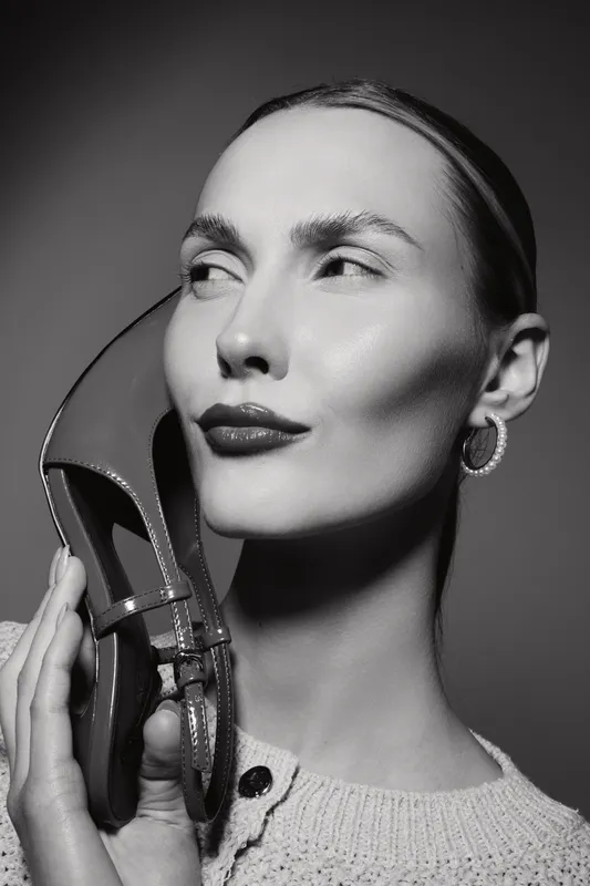 Young white woman with pale skin holds a shoe near her face in a black and white fashion portrait. Concept of style, elegance, and beauty.