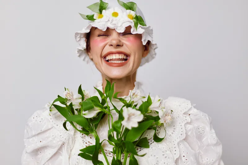 Cheerful young model wearing a floral hat and dress, smiling broadly while holding green flowers against a soft white background, showcasing joy and spring vibes