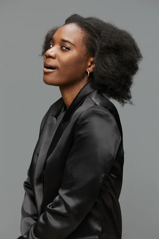 Confident young Black woman in a sleek black blazer, exuding empowerment and joy against a simple gray background, perfect for promoting self esteem and professional themes